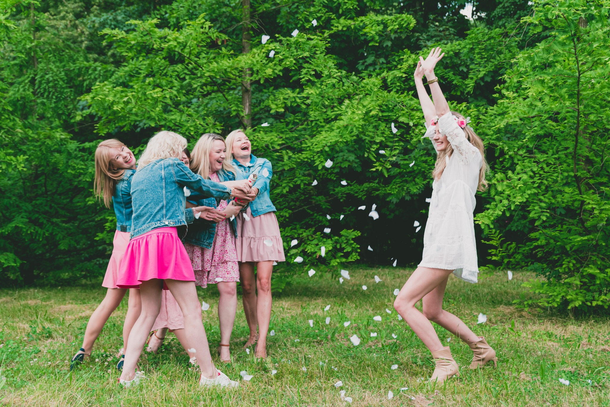 Panna młoda w krótkiej białej sukience i wianku z kwiatów unosi ręce do góry, podczas gdy druhny w jeansowych kurtkach i różowych sukienkach rzucają w nią płatkami kwiatów na tle zielonego lasu.