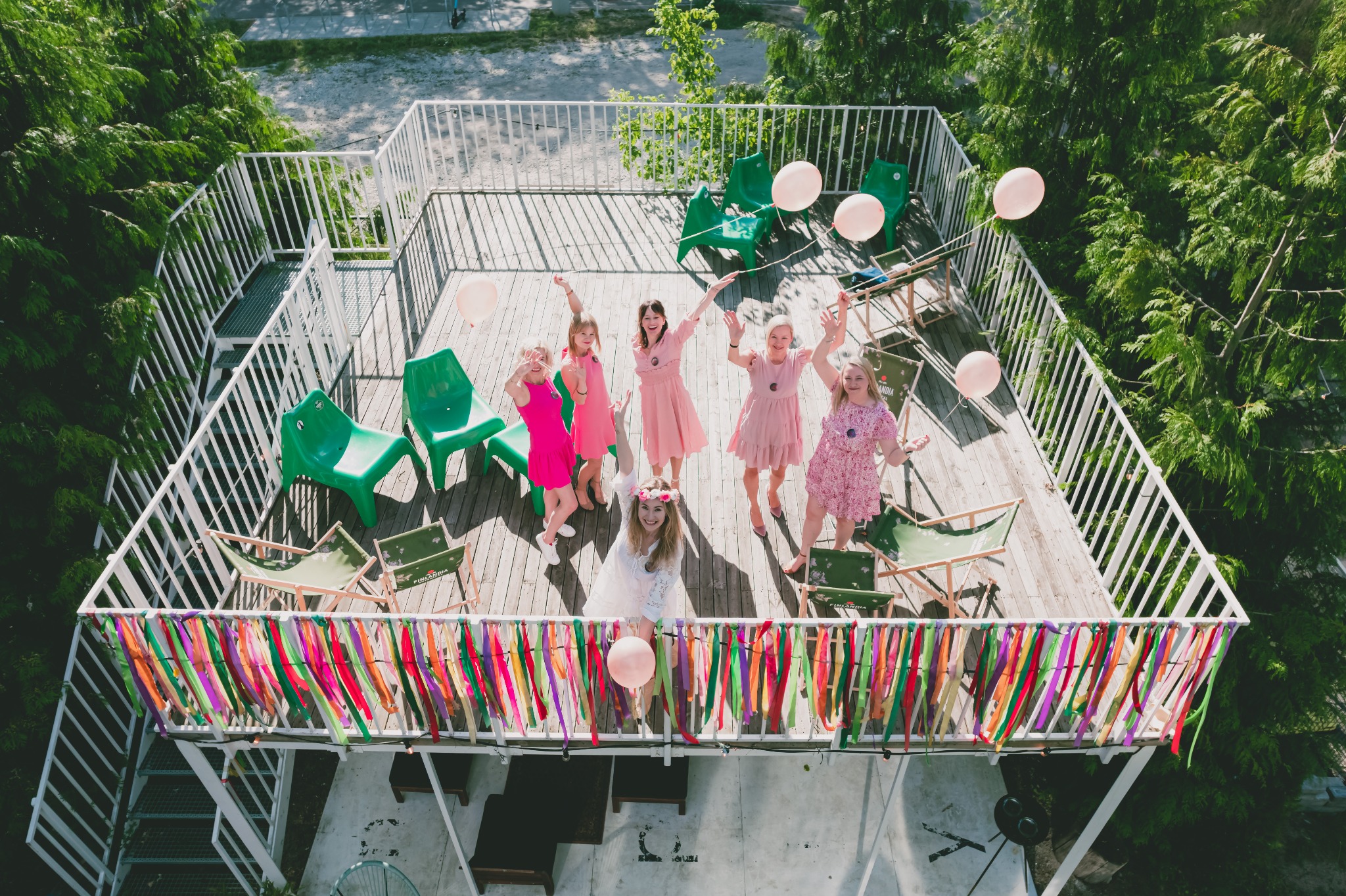 Grupa kobiet w różowych sukienkach i panna młoda w bieli, pozują na drewnianym tarasie z kolorowymi wstążkami na balustradzie, trzymając różowe balony, widok z góry.