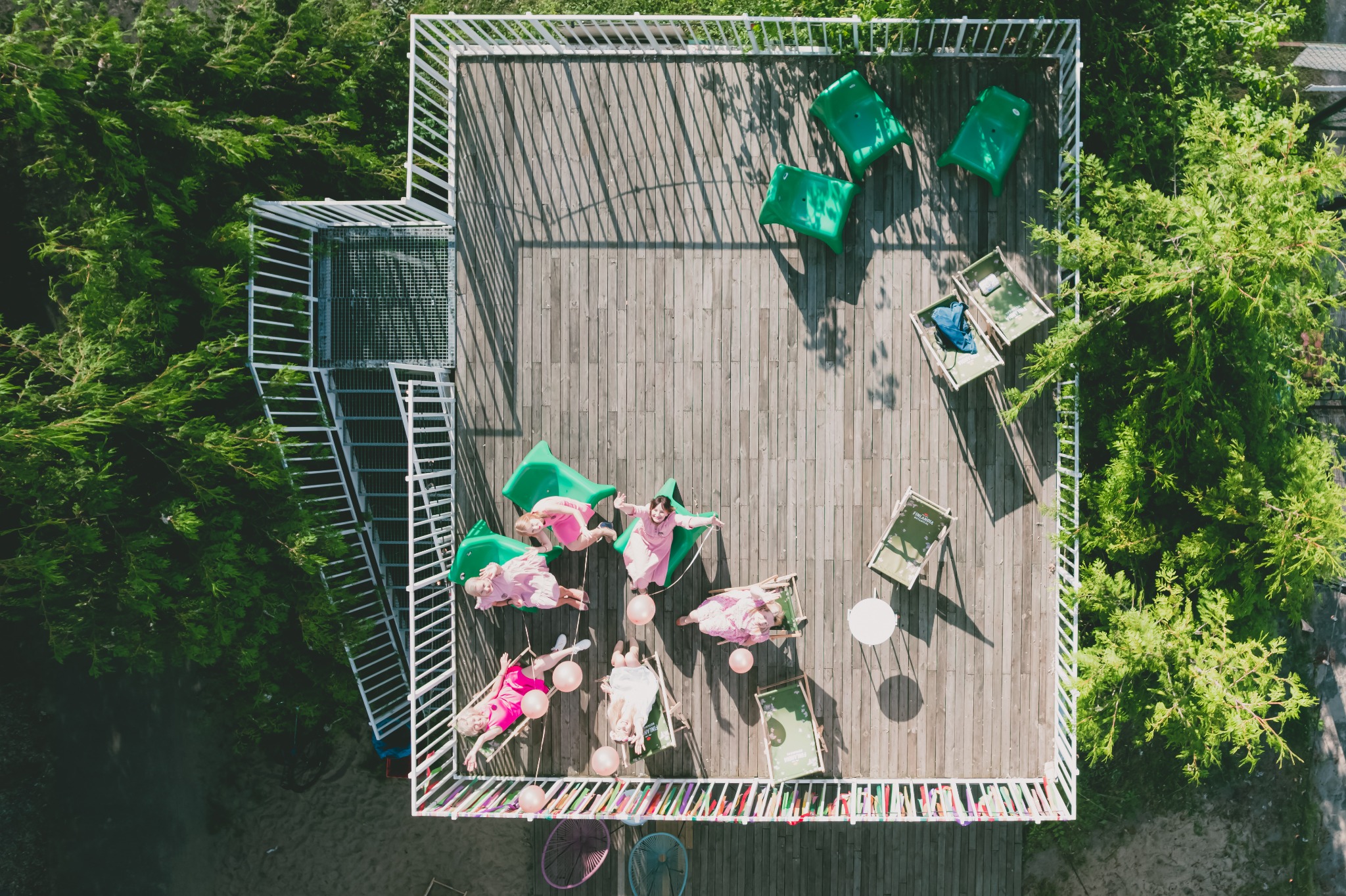 Zdjęcie z lotu ptaka przedstawia grupę kobiet w różowych strojach leżących na leżakach i zielonych krzesłach na drewnianym tarasie otoczonym zielenią, z różowymi balonami, białą balustradą i schodami.