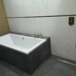 Nowoczesna łazienka z białą wanną obudowaną ciemnym marmurem, złota armatura, odpływ liniowy i listwa dekoracyjna, ściany wyłożone białym marmurem i drewnianymi lamelami.