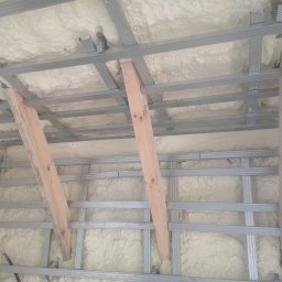 Pro Deco Interior's - Poddasze w trakcie ocieplania pianką poliuretanową, widoczne drewniane podpory i metalowa konstrukcja szkieletowa.