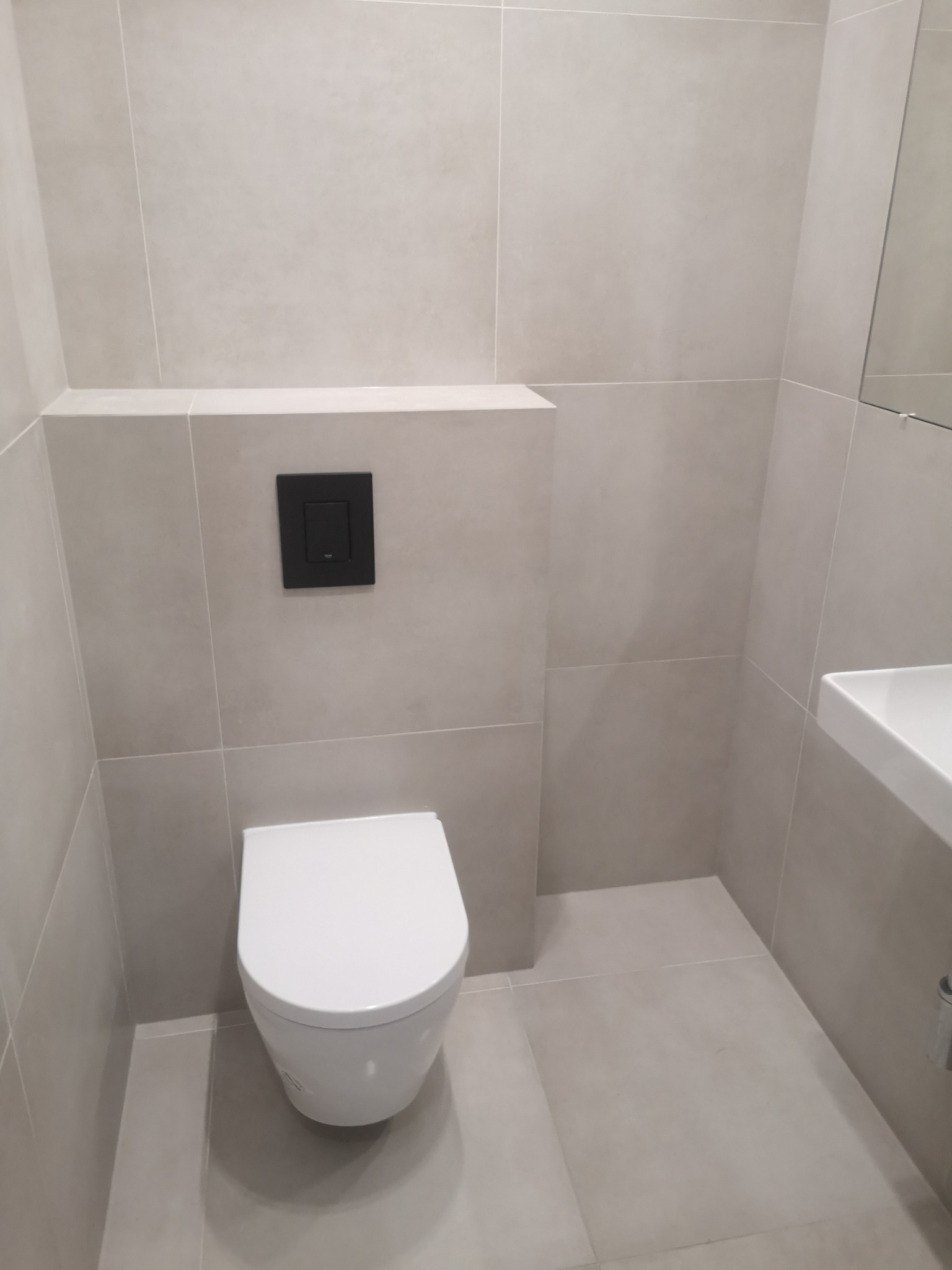 Nowoczesna toaleta podwieszana z czarnym panelem spłukującym, szare płytki ceramiczne na ścianach i podłodze, minimalistyczny design łazienki.