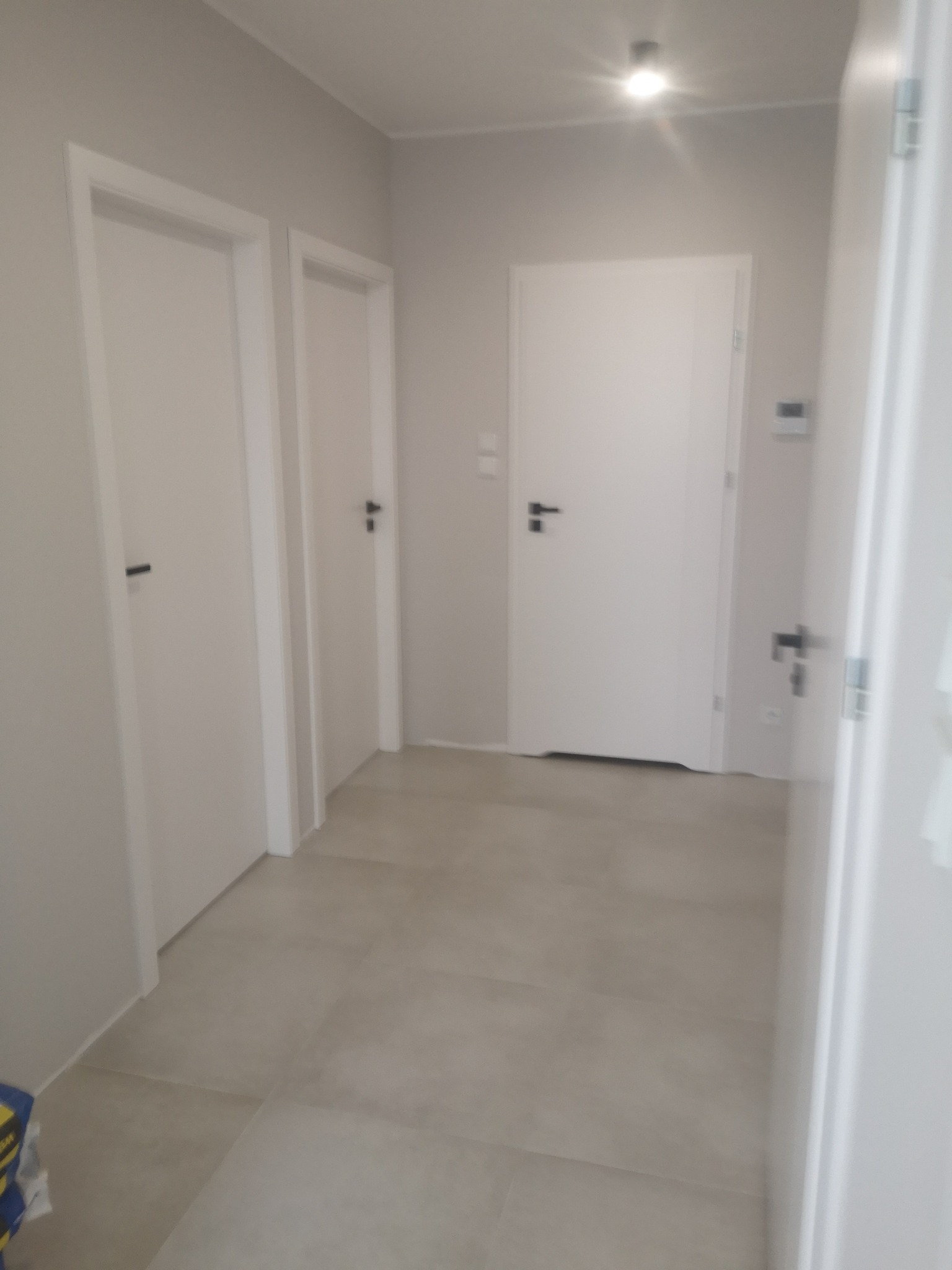 Korytarz w jasnych barwach z trzema białymi drzwiami z czarnymi klamkami, wykończony płytkami w odcieniach beżu.