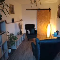 Przytulne wnętrze gabinetu z dwoma ciemnozielonymi fotelami welurowymi, drewnianą podłogą, lampą podłogową z abażurem z naturalnych materiałów, przesuwnymi drzwiami z płyty OSB i dekoracjami...