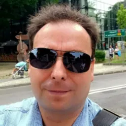 Mężczyzna w okularach przeciwsłonecznych robiący selfie na tle budynku z odbijającym się niebem i rzeźbą kontrabasu oraz znakiem drogowym.