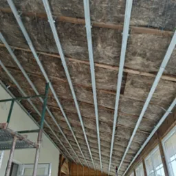 Ocieplenie poddasza wełną mineralną z zamontowanymi metalowymi profilami, widoczne drewniane belki i sznurki mocujące, w tle okna i rusztowanie.