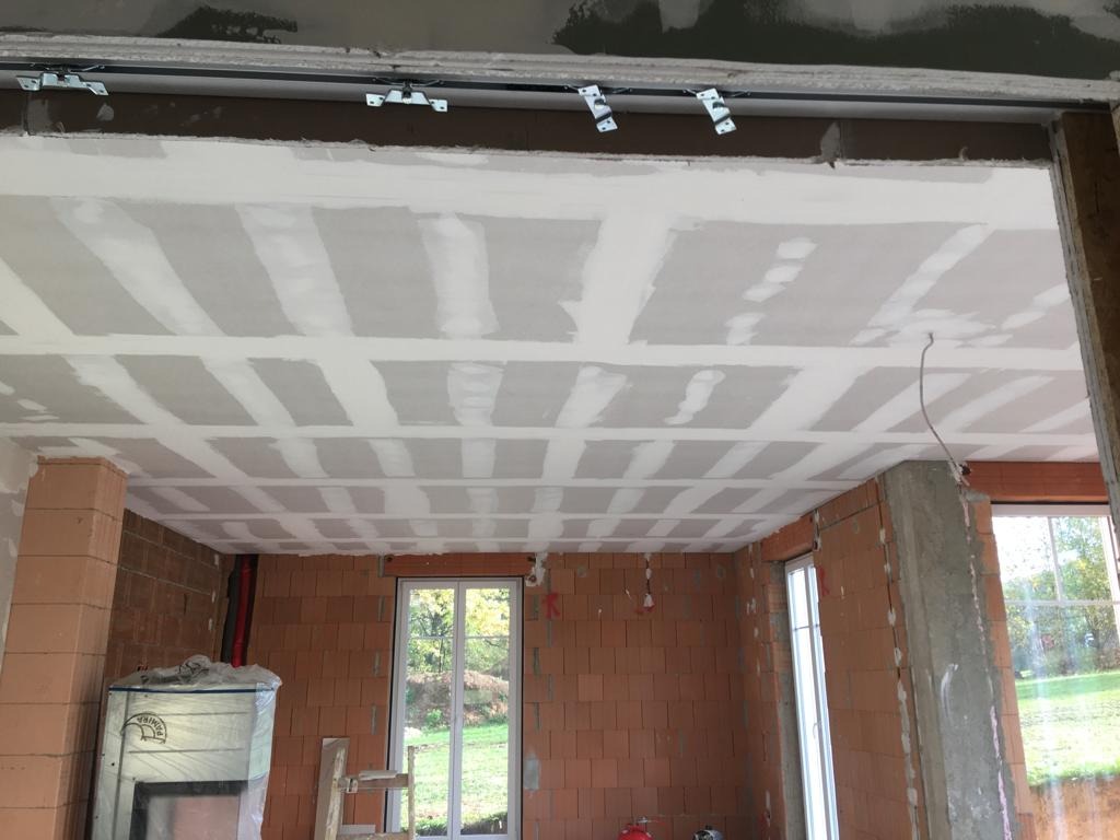 Wnętrze w trakcie prac wykończeniowych: sufit z płyt kartonowo-gipsowych, wstępnie wyszpachlowany, widoczne profile i zawiesia, ściany z cegły dziurawki, okna z widokiem na zieleń.