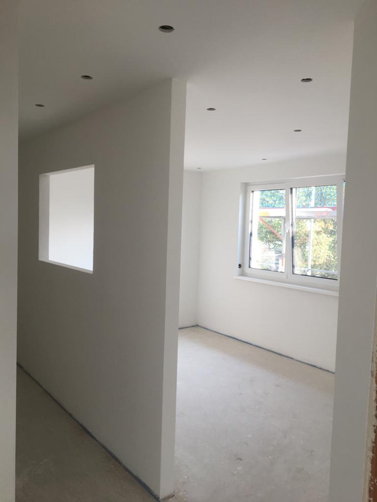 Wnętrze nowo wykończonego, jasnego pomieszczenia z białymi ścianami, widoczne punktowe oświetlenie sufitowe i okno z widokiem na zieleń. Podłoga przygotowana do dalszych prac wykończeniowych.