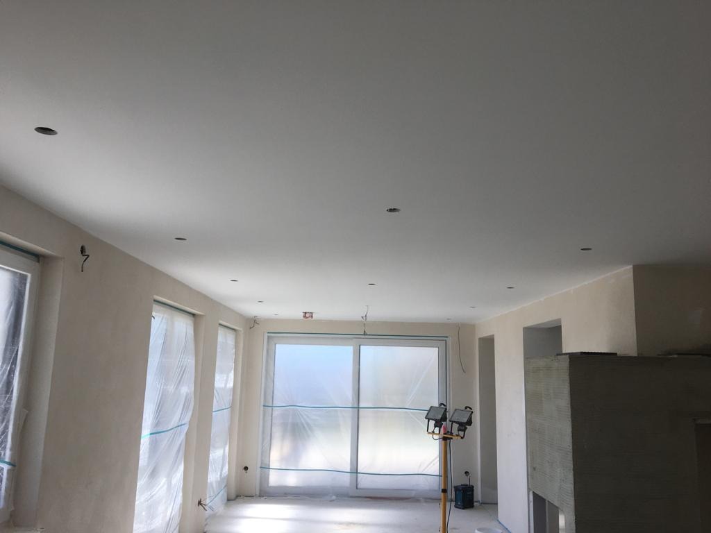 Wnętrze w trakcie remontu: pomalowany sufit z otworami na oświetlenie, okna zabezpieczone folią malarską, widoczne lampy budowlane i płyty karton-gips.
