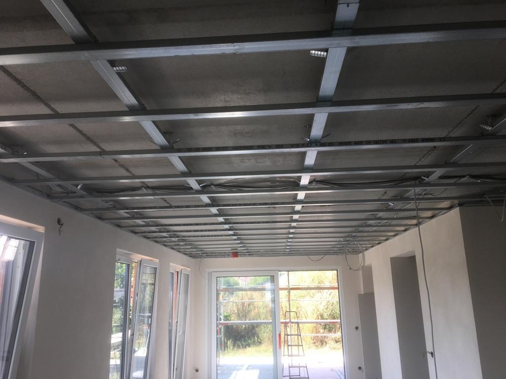 Montaż metalowej konstrukcji podwieszanego sufitu w jasnym pomieszczeniu z widokiem na zewnątrz przez duże okna.