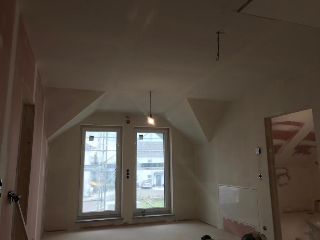 Pokój na poddaszu w trakcie prac wykończeniowych: ściany zagruntowane, fragmenty płyt karton-gips na ścianie, okna dachowe z widokiem na dom sąsiada.