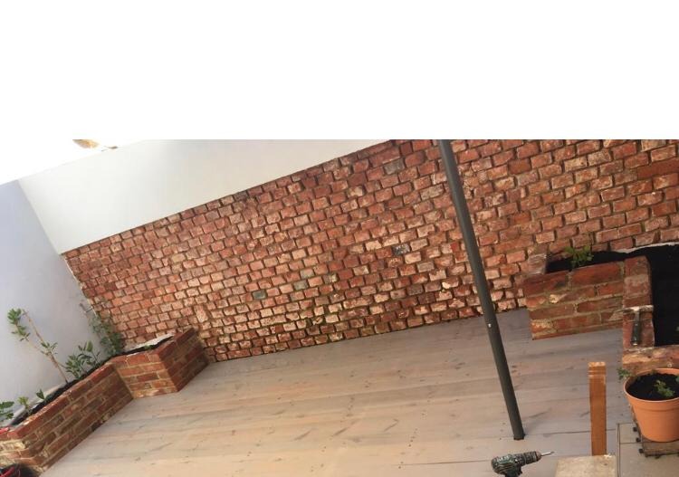 Wykończony taras z drewnianą podłogą, ceglaną ścianą i donicami z roślinami; w rogu widoczna wiertarka.