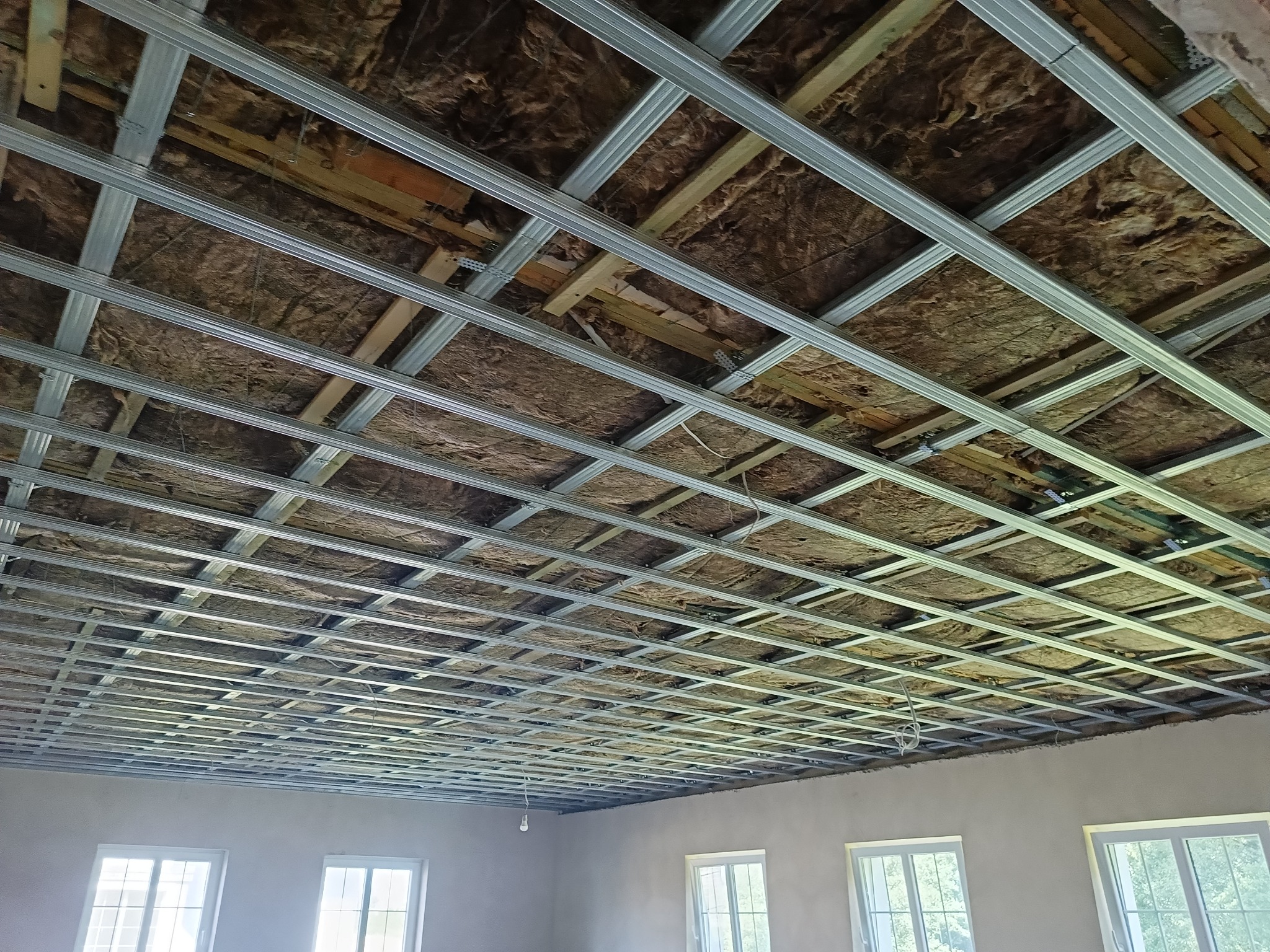 Sufit w trakcie montażu: widoczna konstrukcja metalowa podwieszana, ocieplenie z wełny mineralnej, surowy stan ścian, trzy okna z widokiem na zieleń.