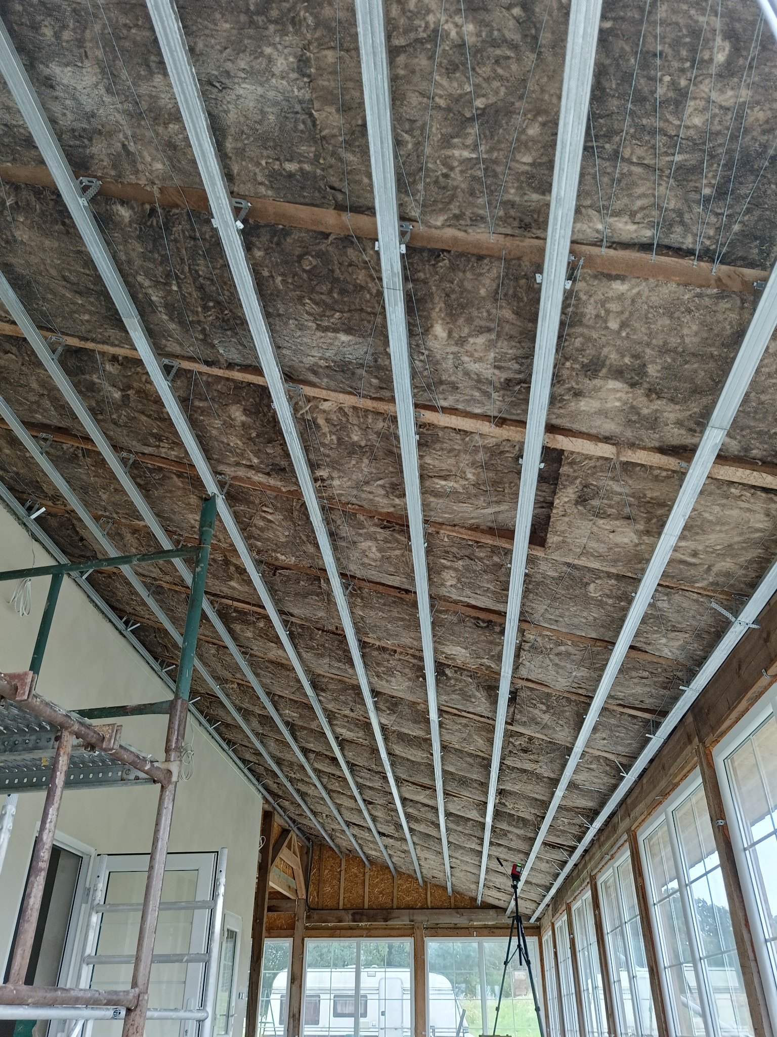 Ocieplenie poddasza wełną mineralną z zamontowanymi metalowymi profilami, widoczne drewniane belki i sznurki mocujące, w tle okna i rusztowanie.