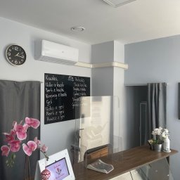 AGood Clima Aleksander Gorgolewski - Wnętrze lokalu gastronomicznego z zamontowaną klimatyzacją ścienną marki ANDE, widoczny zegar, tablica z menu, osłona z pleksi na ladzie oraz dekoracje roślinne.