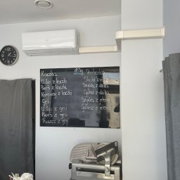 AGood Clima Aleksander Gorgolewski - Wnętrze lokalu gastronomicznego z klimatyzatorem ściennym marki ANDE, zegarem analogowym, czarną tablicą z menu wypisanym kredą (kaczka, gęś, perliczka, dzik, jeleń) oraz krajalnicą do wędlin...