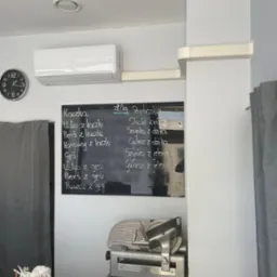 Wnętrze lokalu gastronomicznego z klimatyzatorem ściennym marki ANDE, zegarem analogowym, czarną tablicą z menu wypisanym kredą (kaczka, gęś, perliczka, dzik, jeleń) oraz krajalnicą do wędlin...