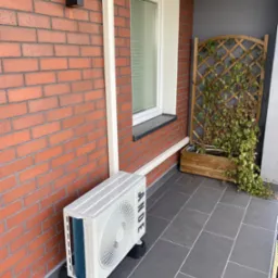 Biała jednostka zewnętrzna klimatyzacji, zamontowana na czarnych podstawkach, stoi na szarym balkonie przy ceglanej ścianie budynku. Widoczna biała rura odprowadzająca skropliny. W tle drewniana...