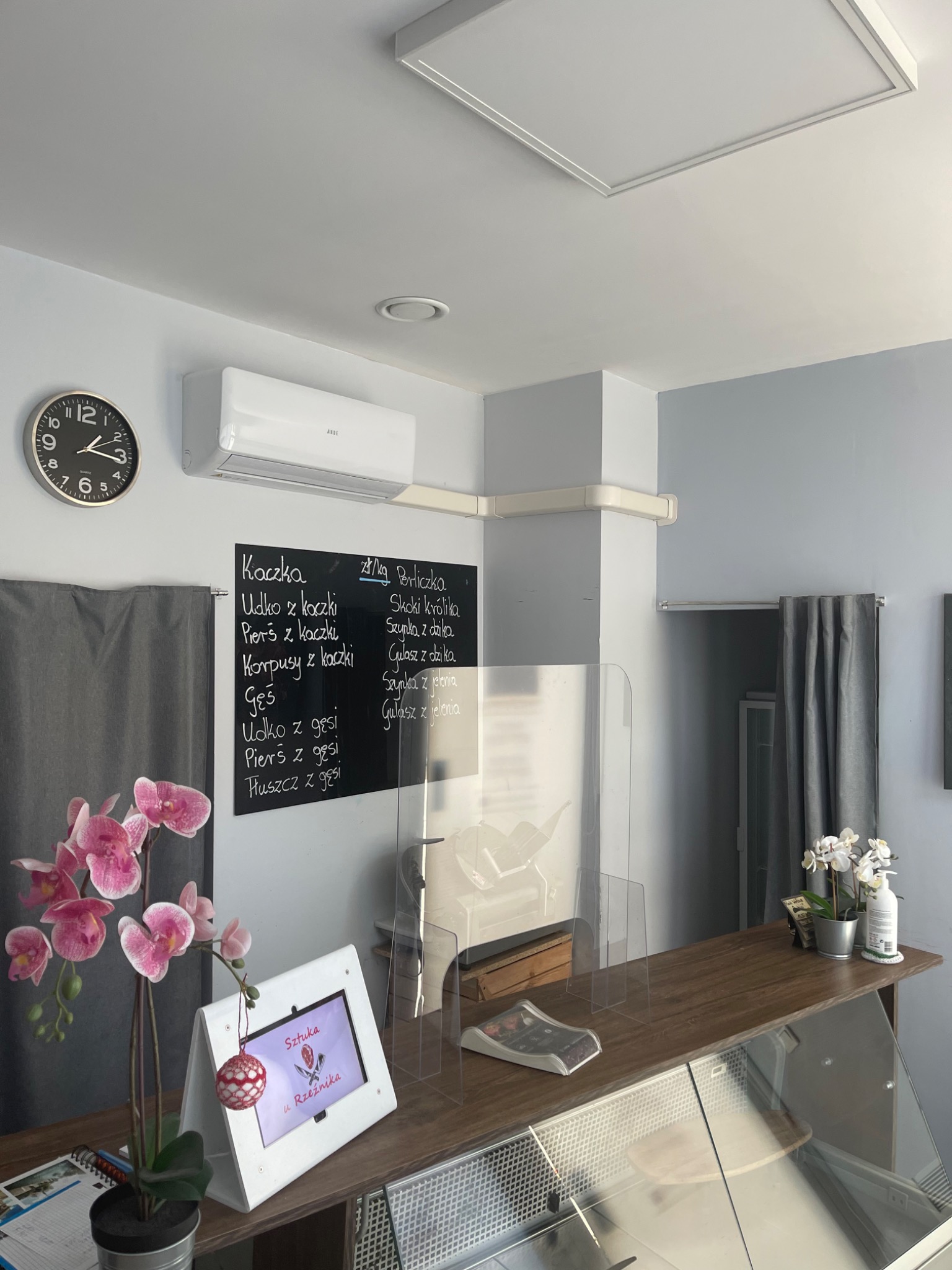 Wnętrze lokalu gastronomicznego z zamontowaną klimatyzacją ścienną marki ANDE, widoczny zegar, tablica z menu, osłona z pleksi na ladzie oraz dekoracje roślinne.