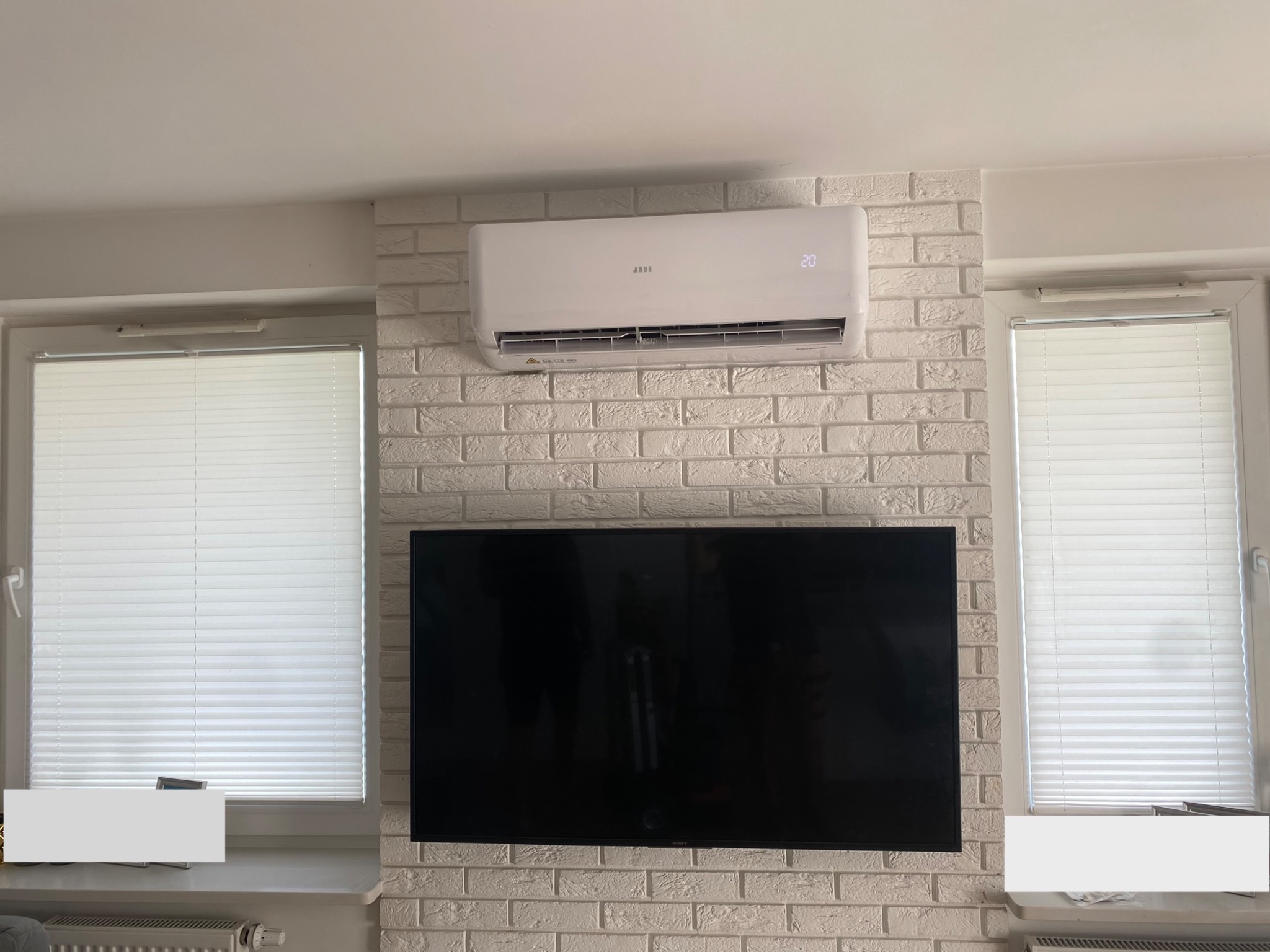 Biała jednostka klimatyzacyjna marki ANDE zamontowana na ścianie z białej cegły w pokoju z dwoma oknami z żaluzjami i telewizorem.