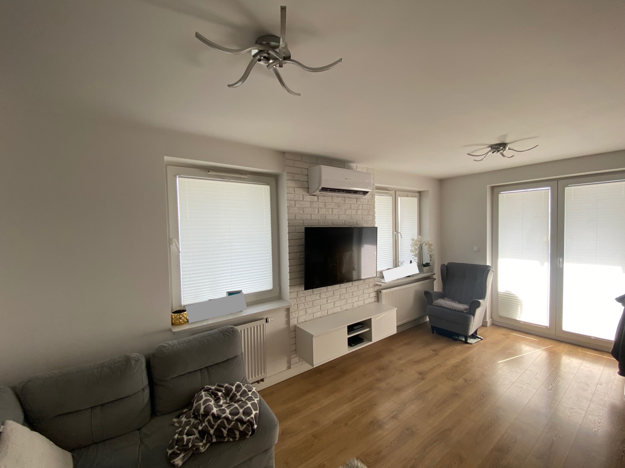 Jasne wnętrze salonu z klimatyzatorem ściennym zamontowanym nad telewizorem na ścianie z białej cegły, szara sofa i fotel, drewniana podłoga.
