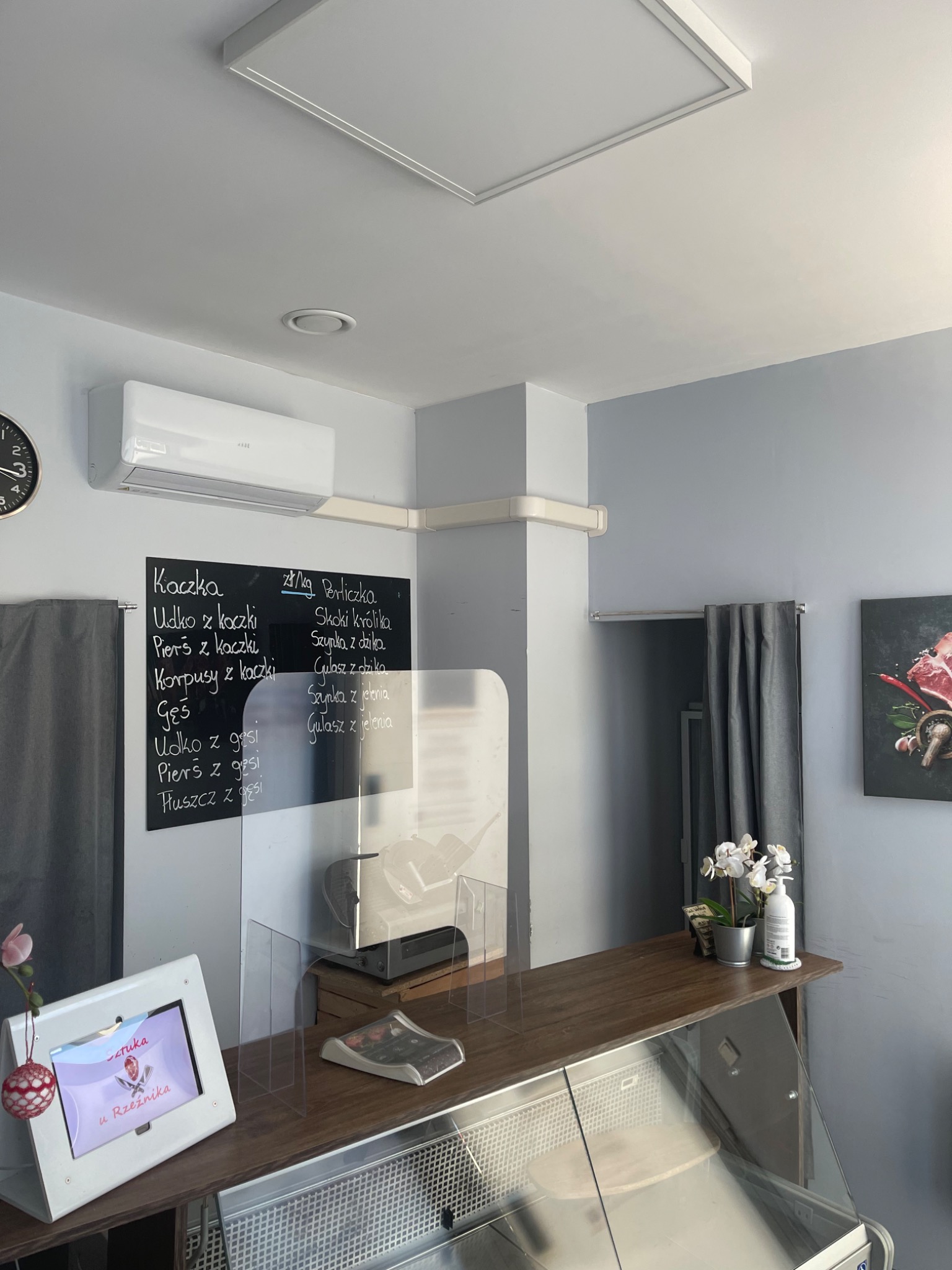 Wnętrze sklepu mięsnego z zamontowaną klimatyzacją typu split na ścianie, widoczna tablica z menu, lada chłodnicza i elementy dekoracyjne.