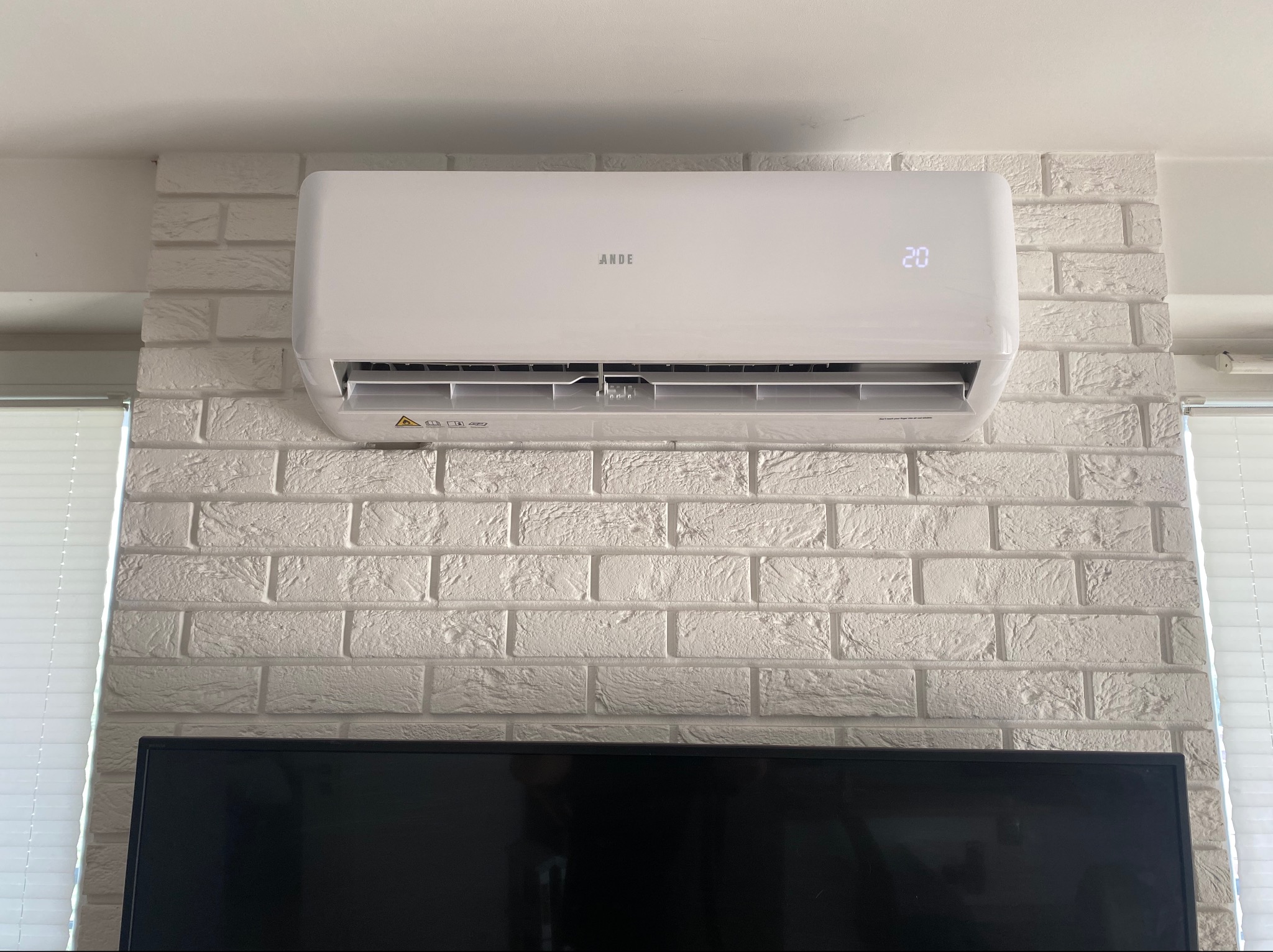 Biały klimatyzator marki ARDE zamontowany na ścianie z dekoracyjnej białej cegły, widoczny wyświetlacz z temperaturą 20 stopni Celsjusza, pod nim fragment czarnego telewizora.