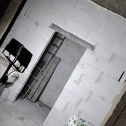 Wnętrze w trakcie budowy: ściany z białych bloczków, widoczne zaprawy, drabina oparta o ścianę, reflektor oświetlający przestrzeń i worki z zaprawą murarską ułożone na palecie.