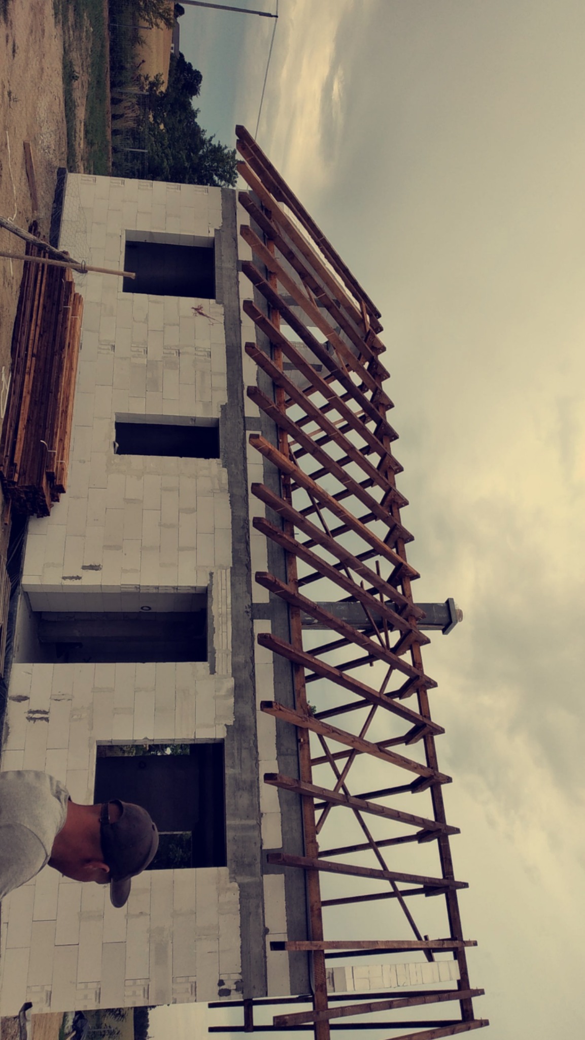 Wznoszenie dwupiętrowego domu z białych bloczków, widoczny szkielet dachu z drewnianych belek i fragment sylwetki pracownika w szarej bluzie i czapce z daszkiem.