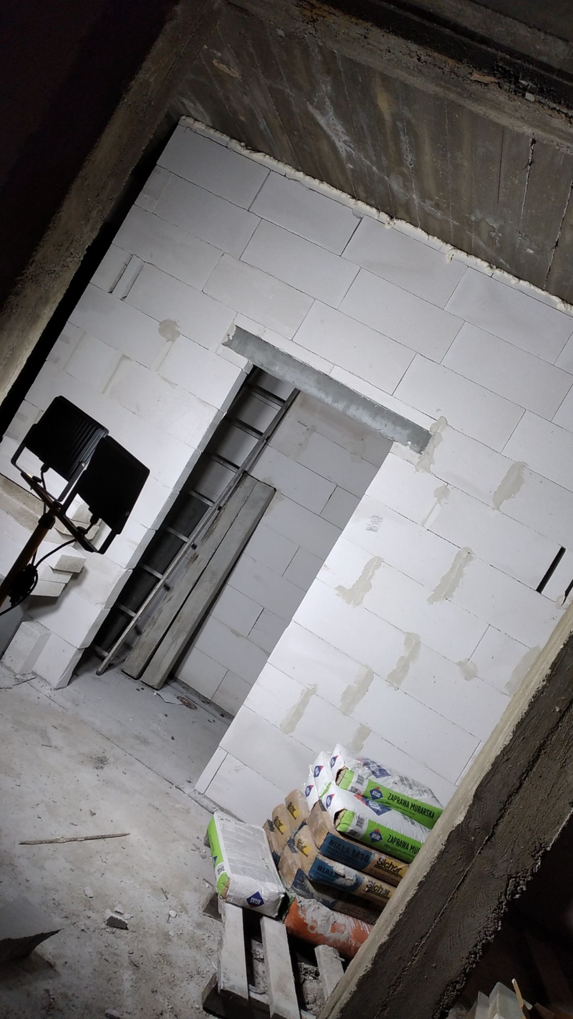 Wnętrze w trakcie budowy: ściany z białych bloczków, widoczne zaprawy, drabina oparta o ścianę, reflektor oświetlający przestrzeń i worki z zaprawą murarską ułożone na palecie.