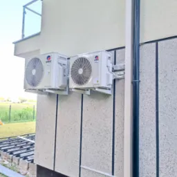 Dwa białe, identyczne klimatyzatory marki Gree zamontowane na elewacji budynku z widocznymi przewodami i elementami instalacji, na tle trawnika i nieba.