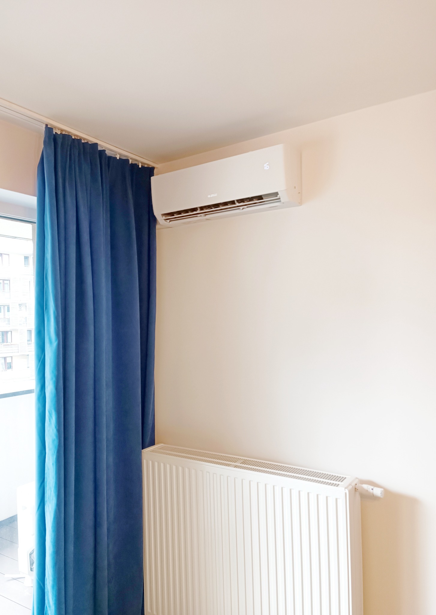 Biały klimatyzator ścienny zamontowany nad białym grzejnikiem, obok okna z zasłoniętymi niebieskimi zasłonami w jasnym pokoju.