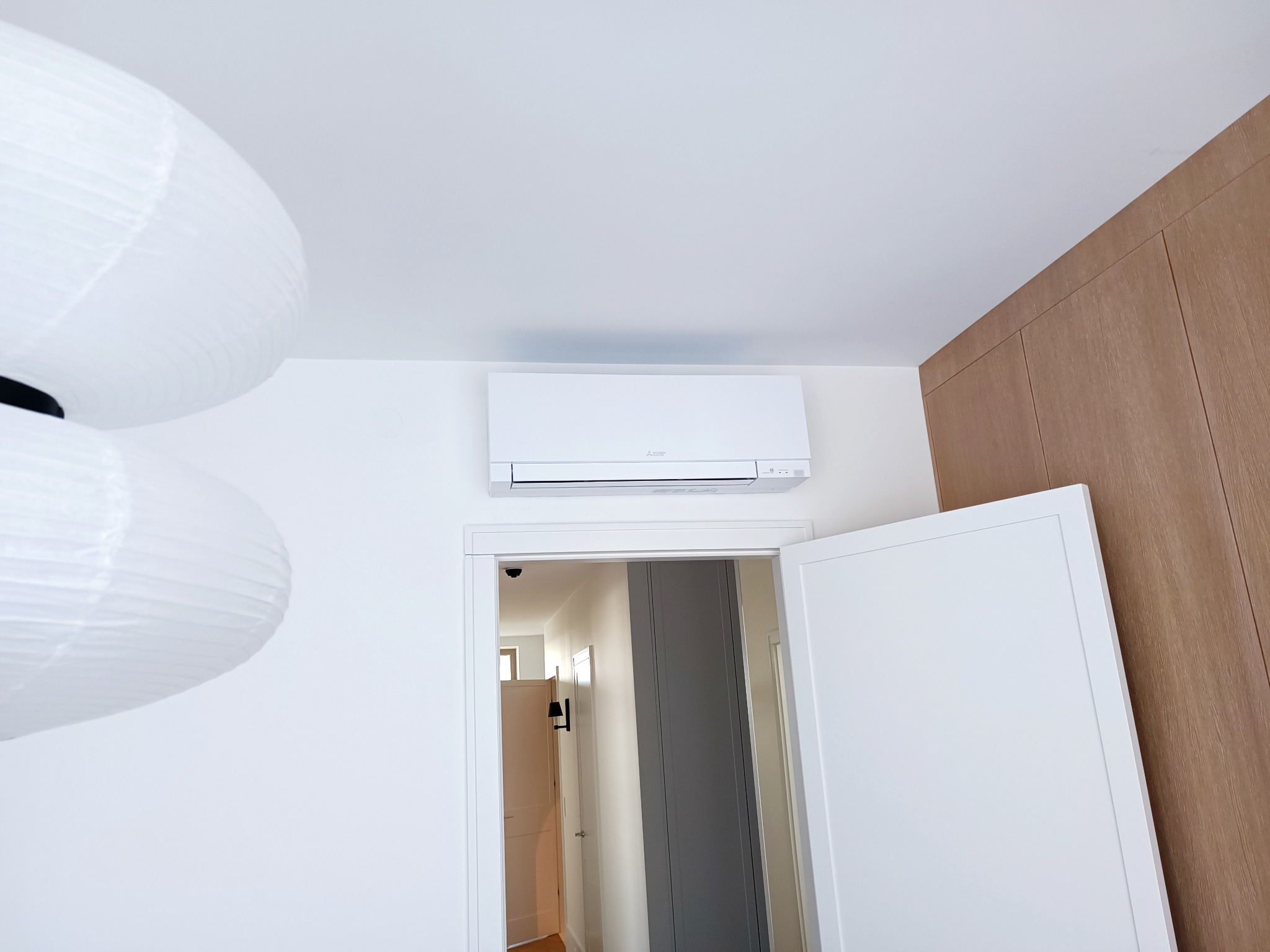 Biały klimatyzator Mitsubishi zamontowany nad otwartymi drzwiami w jasnym wnętrzu, widoczna lampa papierowa i drewniana szafa w zabudowie.