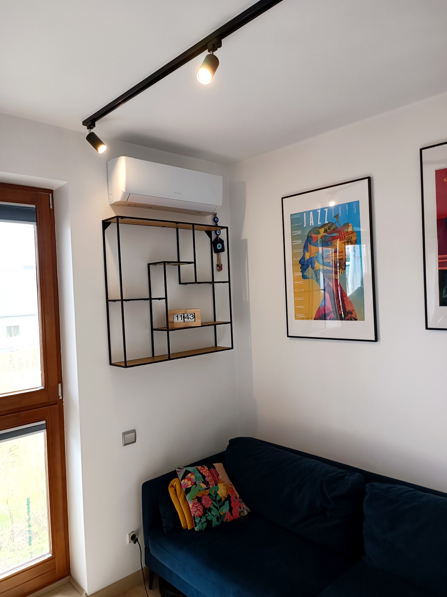 Biała jednostka klimatyzacyjna zamontowana na ścianie nad czarną, geometryczną półką w nowoczesnym salonie z niebieską sofą i artystycznymi plakatami.