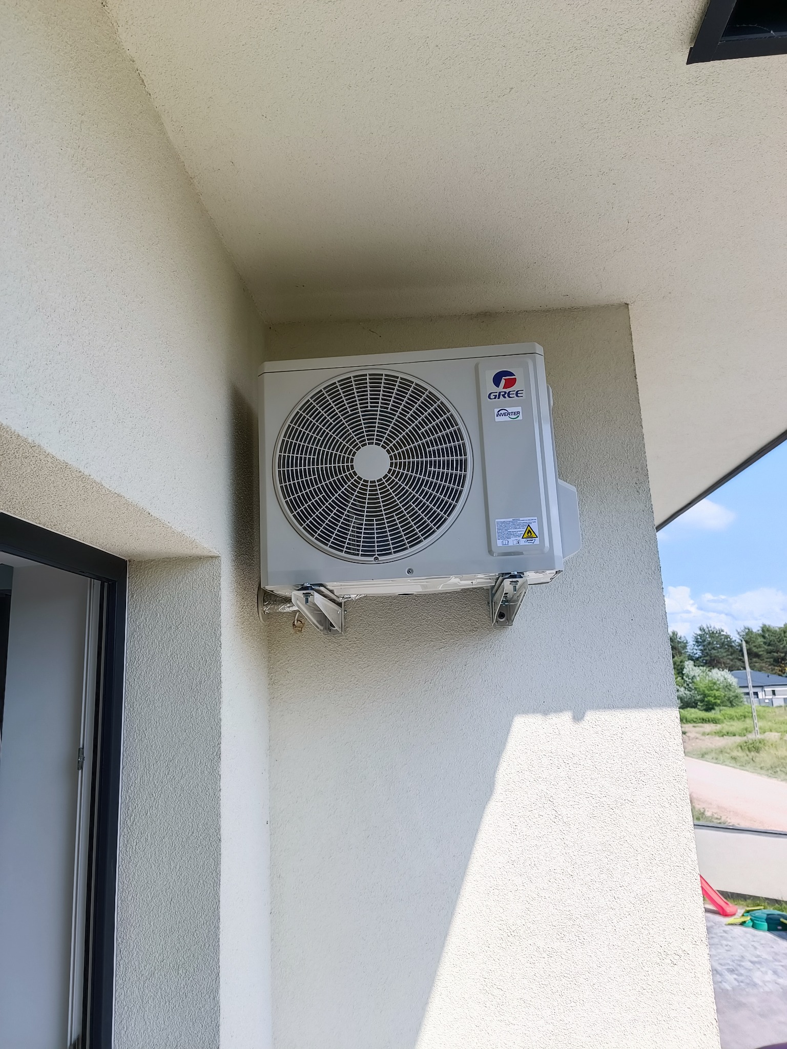 Zewnętrzna jednostka klimatyzacji Gree Inverter zamontowana na ścianie budynku z elewacją o fakturze baranka, widoczne uchwyty montażowe i fragment okna z czarną ramą.