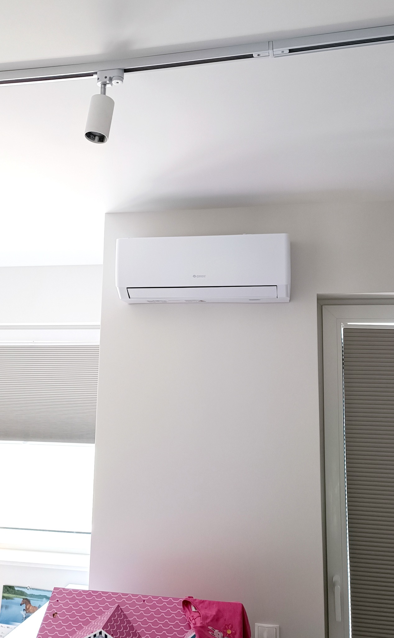 Biały klimatyzator marki Gree zamontowany na ścianie w jasnym pokoju, widoczne szyny oświetleniowe sufitowe z reflektorem, fragment okna z żaluzjami plisowanymi oraz dziecięce zabawki w dolnej...