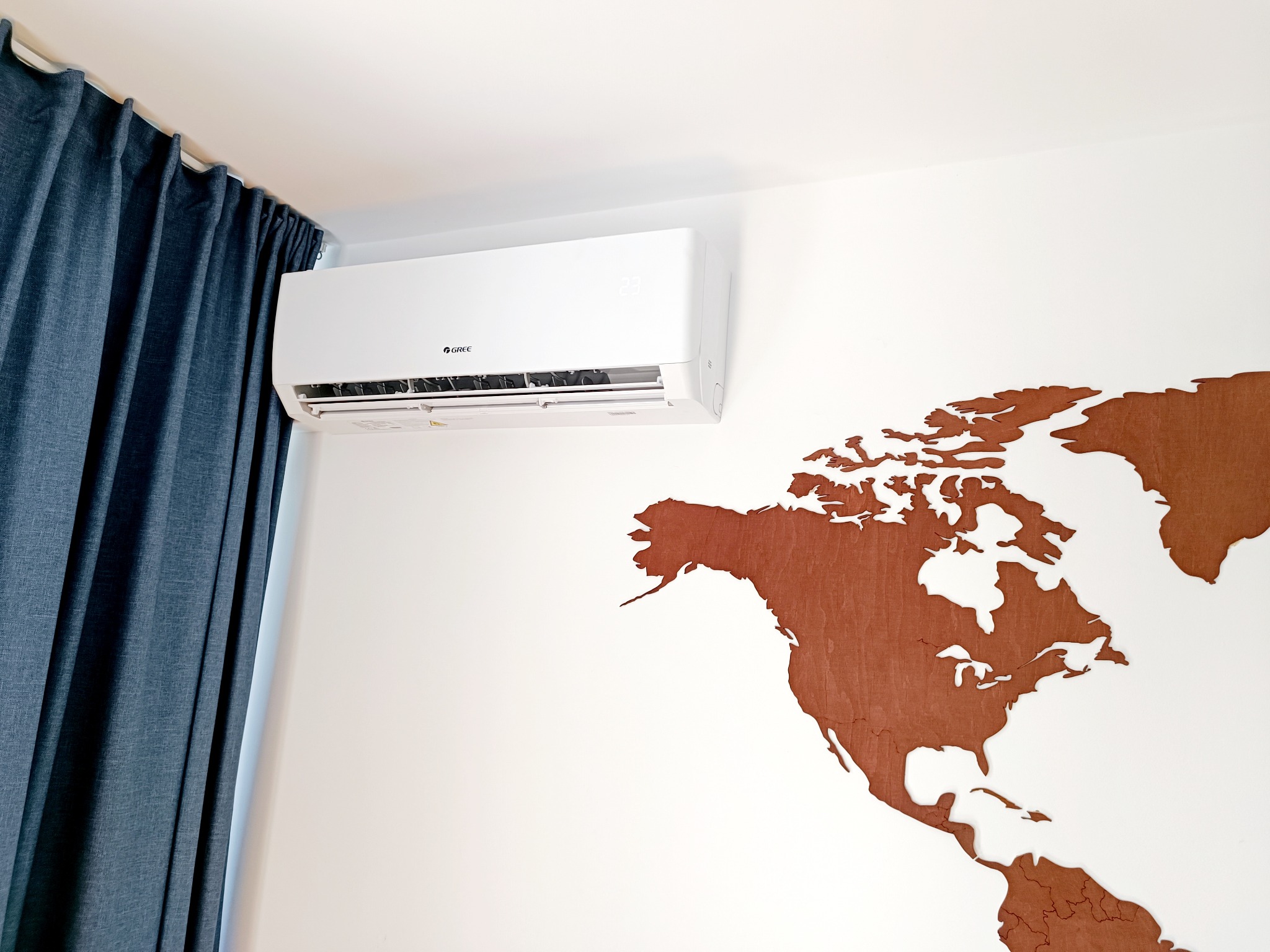 Biały klimatyzator marki Gree zamontowany na ścianie z drewnianą mapą świata, obok zasłoniętego okna.