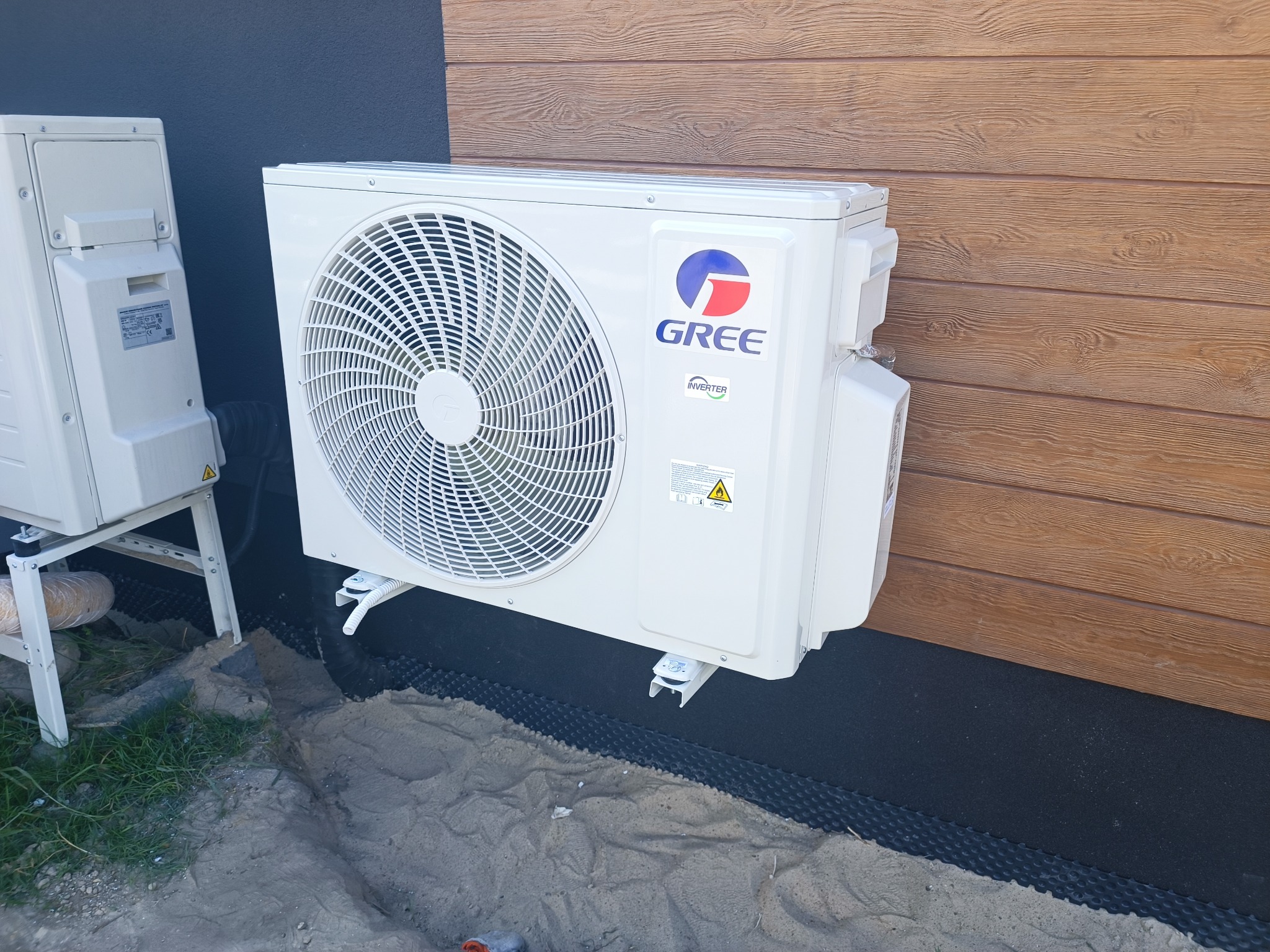Biała jednostka zewnętrzna klimatyzatora marki Gree zamontowana na elewacji budynku z drewnianą okładziną, widoczna również druga jednostka na wspornikach.