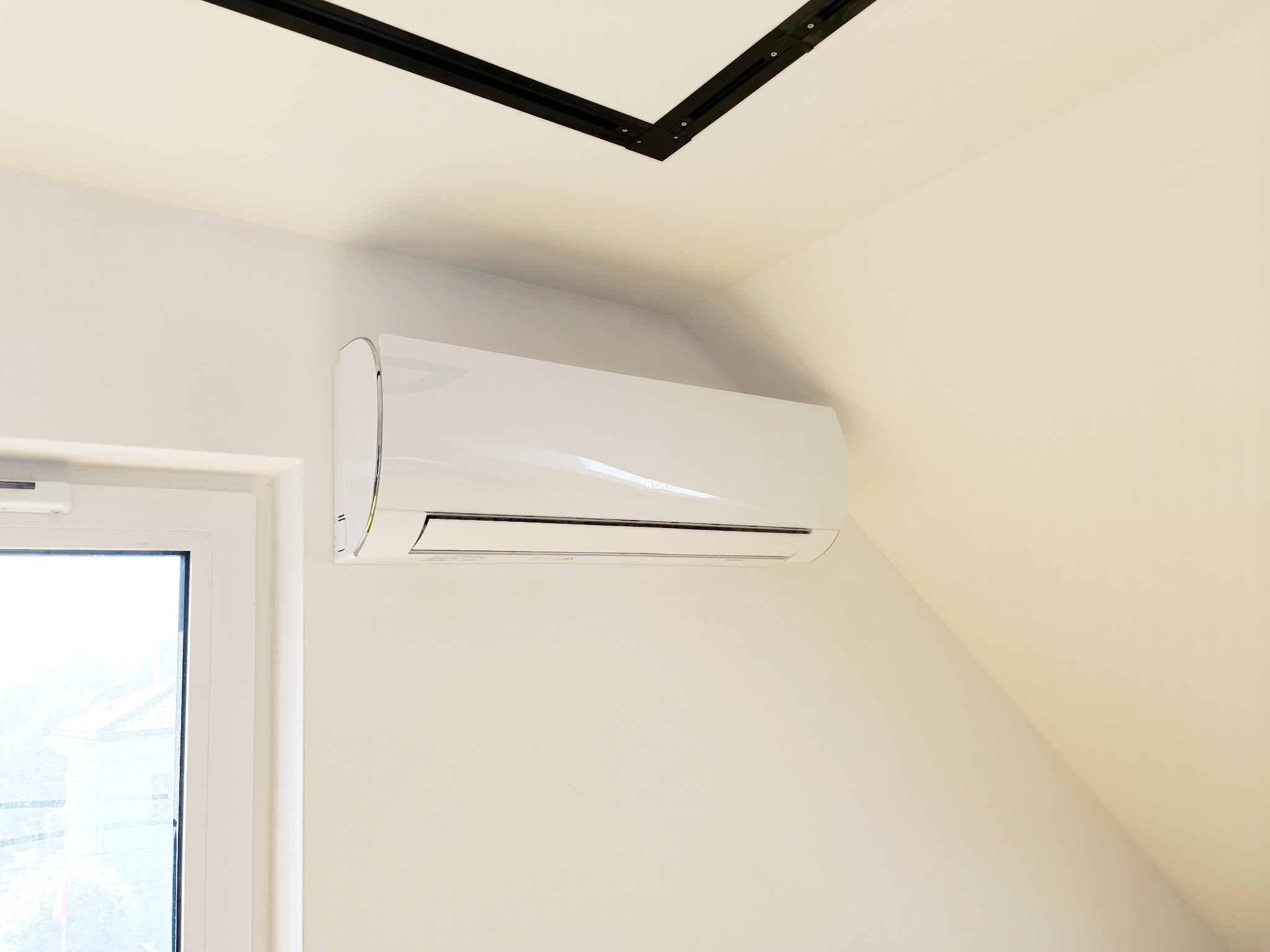 Biały klimatyzator ścienny zamontowany pod skosem poddasza, obok okna z widokiem na zewnątrz.