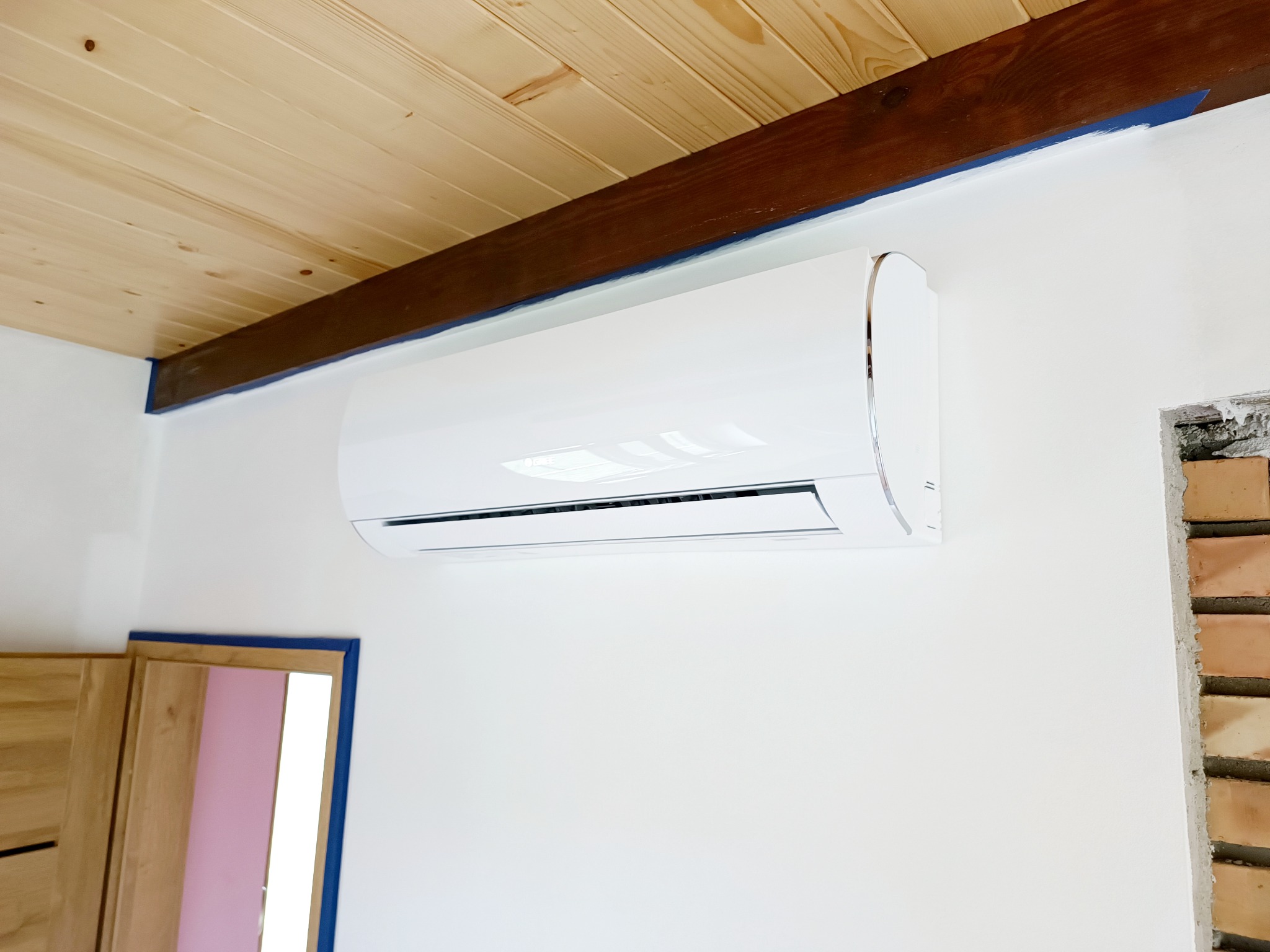 Biały klimatyzator ścienny zamontowany w pomieszczeniu z drewnianym sufitem i fragmentem ściany z odsłoniętą cegłą w otworze okiennym.