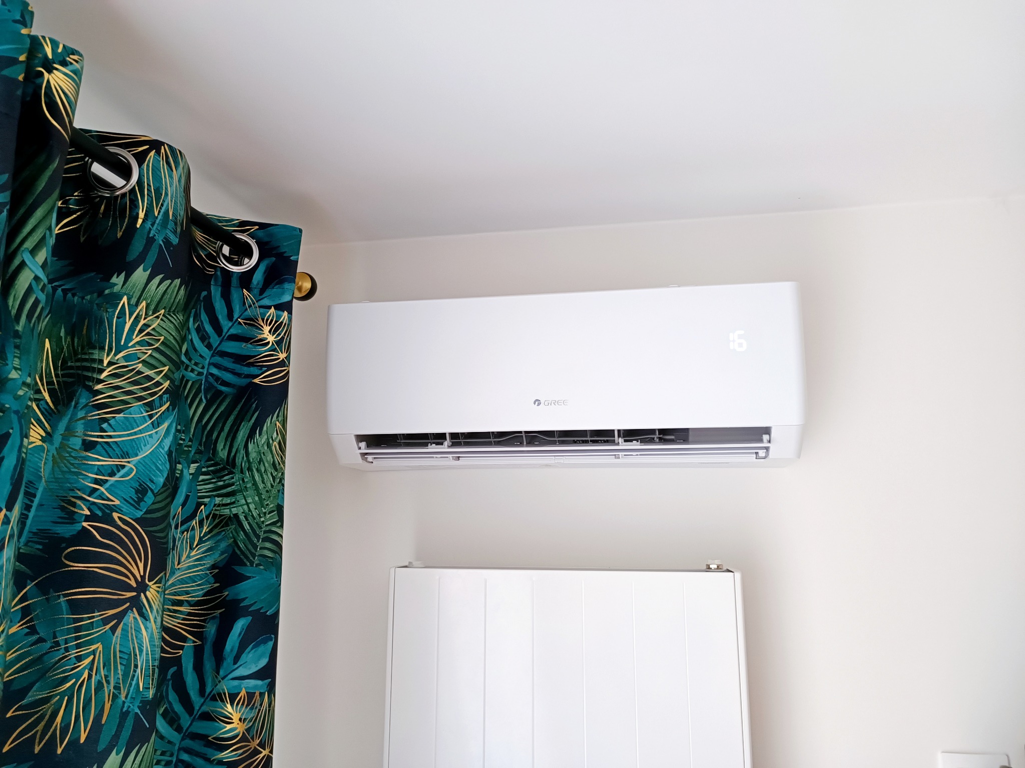 Biały klimatyzator marki Gree zamontowany na ścianie w pokoju z dekoracyjnymi, zielono-złotymi zasłonami w motywy roślinne i białym grzejnikiem panelowym pod spodem.