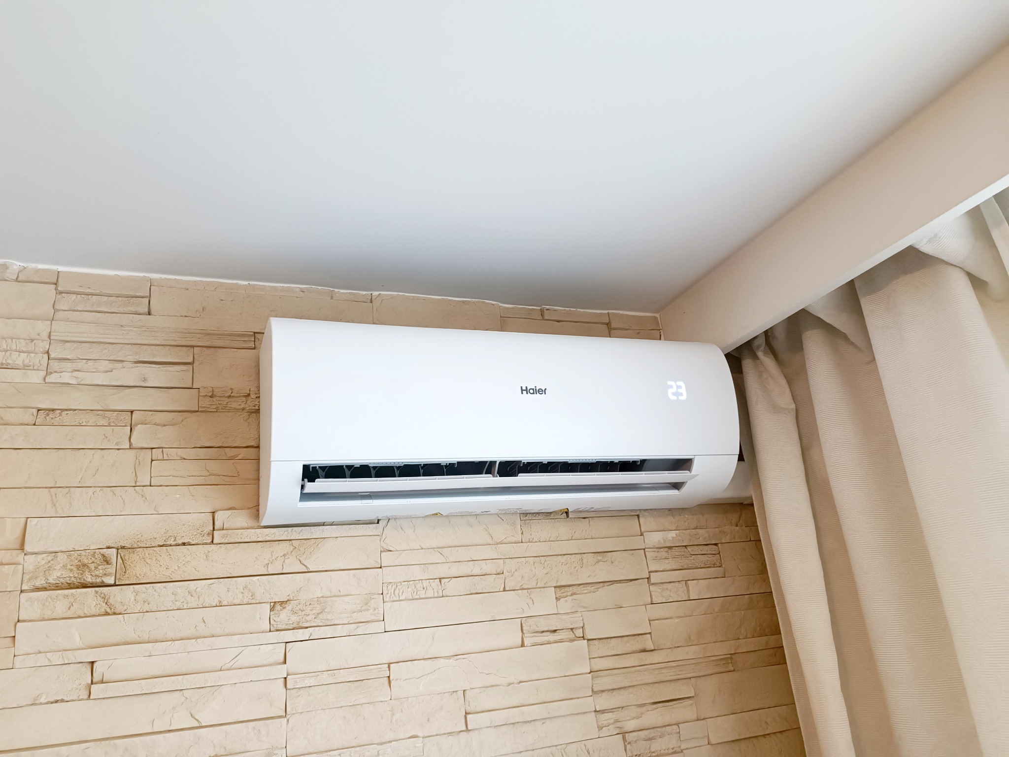 Biały klimatyzator Haier zamontowany na ścianie wyłożonej dekoracyjną cegłą, z wyświetlaczem wskazującym 23 stopnie Celsjusza, obok zwisająca beżowa zasłona.