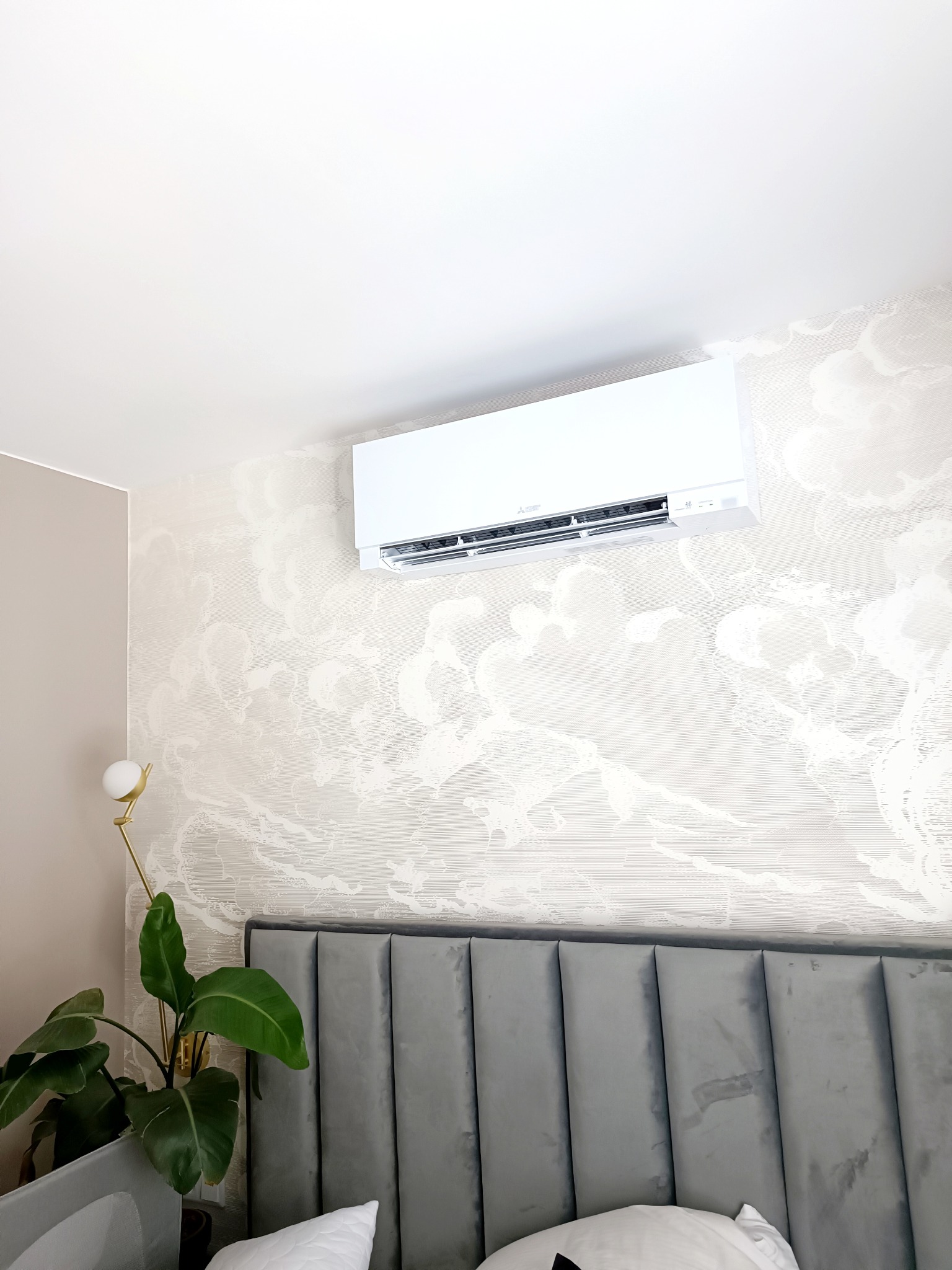 Biały klimatyzator ścienny zamontowany nad wezgłowiem łóżka w sypialni z tapetą w subtelne wzory, oświetlony złotą lampą podłogową obok doniczkowej rośliny.