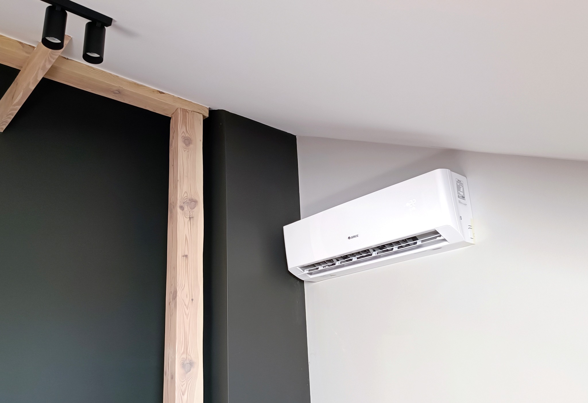 Biały klimatyzator marki Gree zamontowany na jasnej ścianie w nowoczesnym wnętrzu z drewnianymi elementami konstrukcyjnymi i czarną ścianą w tle.