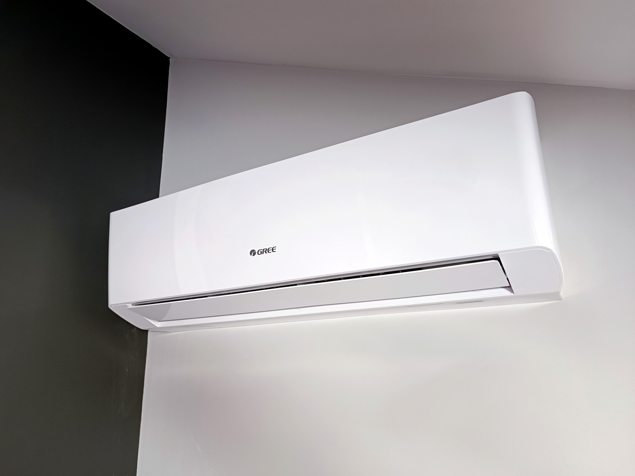 Biała jednostka klimatyzacyjna marki Gree zamontowana na ścianie pomalowanej częściowo na biało, a częściowo na ciemnoszaro.