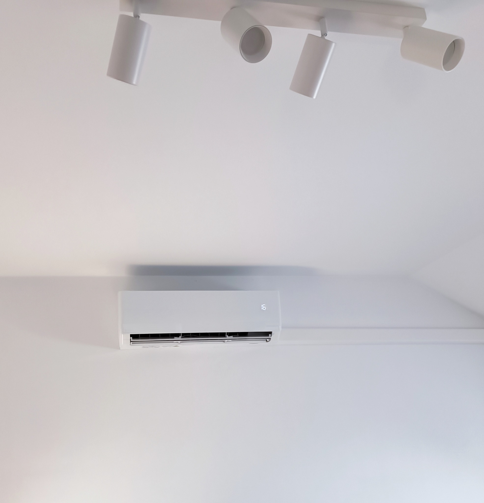 Biała jednostka klimatyzacji zamontowana na białej ścianie pod skośnym sufitem, oświetlona przez reflektory sufitowe