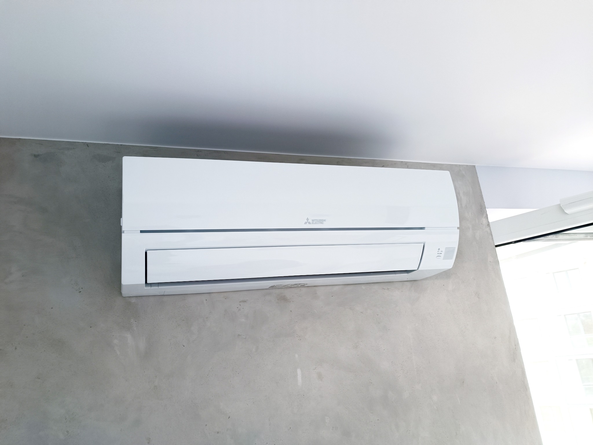 Biała jednostka klimatyzacji Mitsubishi Electric zamontowana na szarej, betonowej ścianie w pomieszczeniu w Grodzisku Mazowieckim. Widoczne okno w tle.