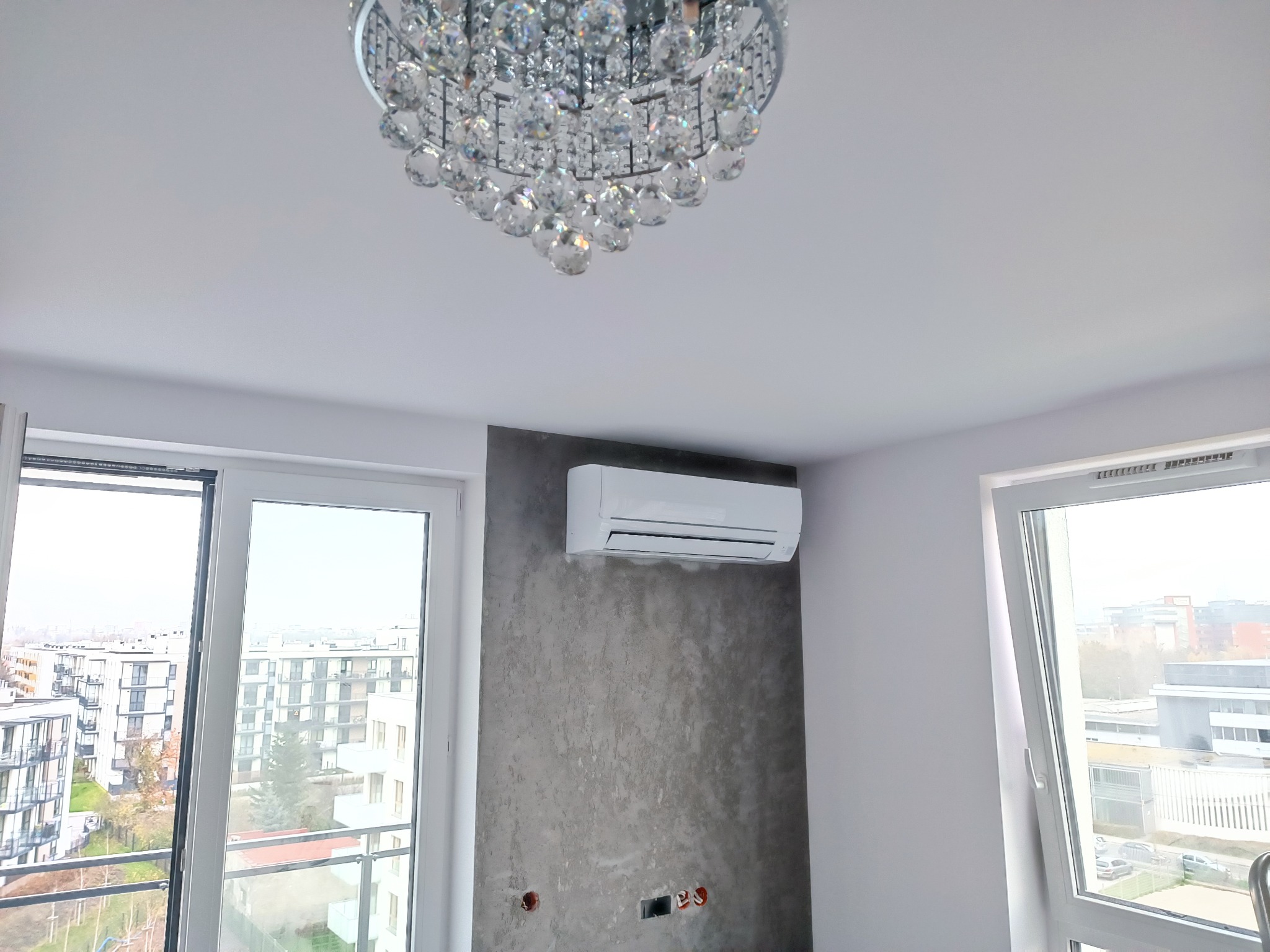 Biały klimatyzator ścienny zamontowany na szarej, teksturowanej ścianie w nowoczesnym wnętrzu z dużym, kryształowym żyrandolem i widokiem na miasto przez okno.