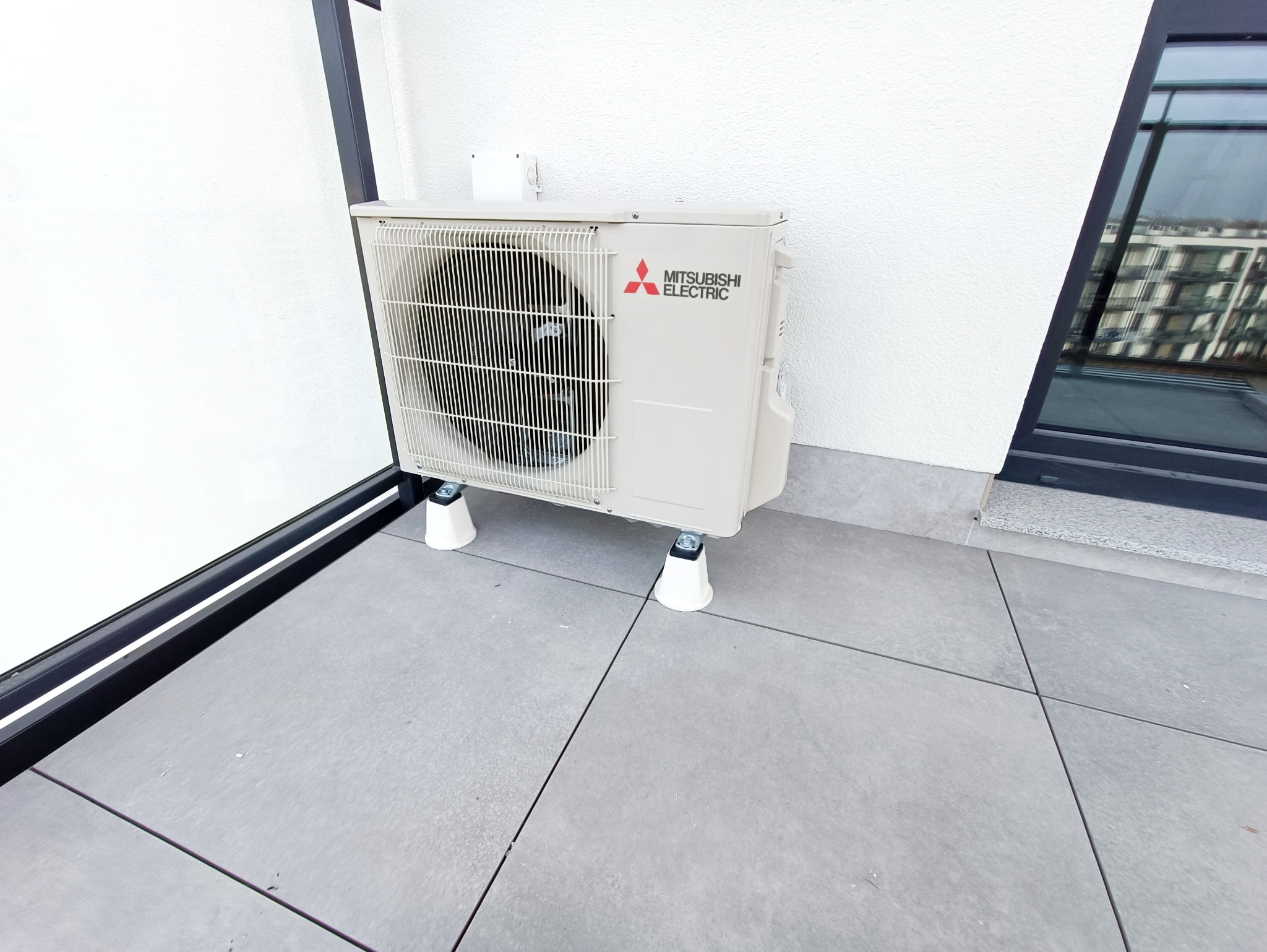 Zewnętrzna jednostka klimatyzacji Mitsubishi Electric na balkonie, zamontowana na białych podkładkach antywibracyjnych, widok z boku.