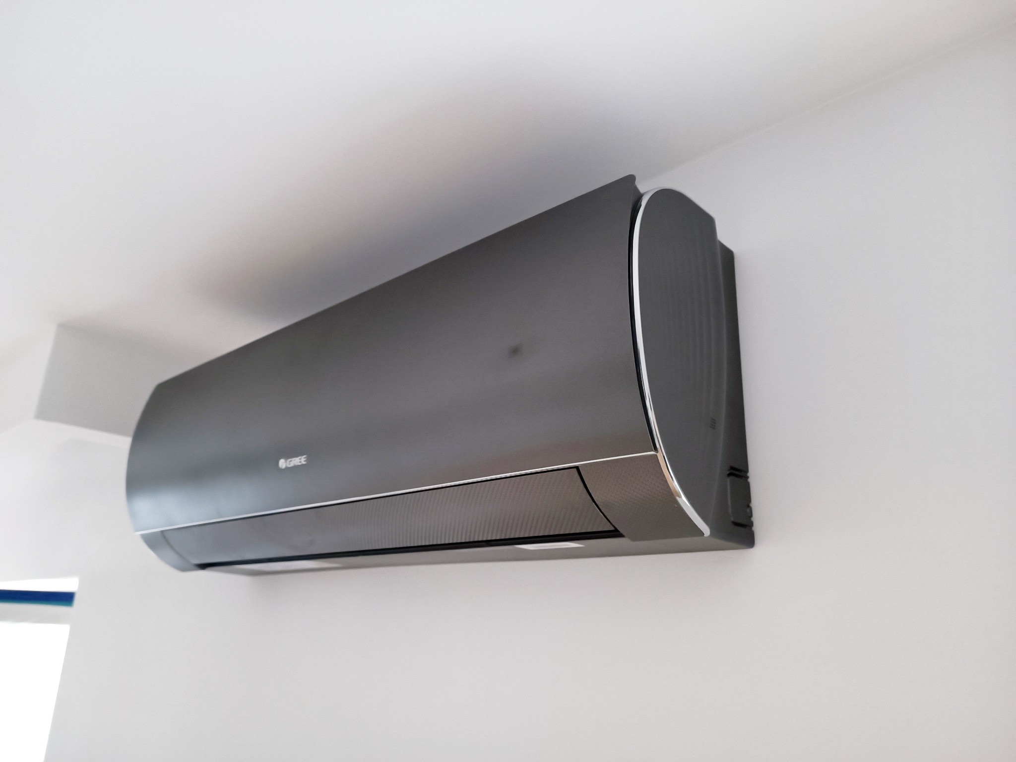 Czarny klimatyzator Gree zamontowany na białej ścianie, widoczny z dołu, z delikatnym odbiciem światła na obudowie.